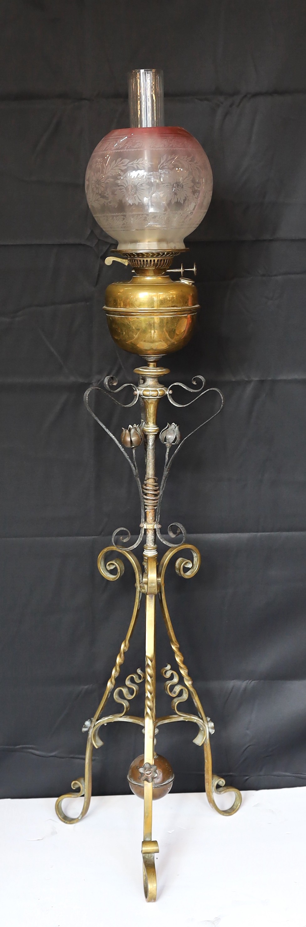A late Victorian Arts and Crafts wrought iron and brass miniature oil lamp standard, in the manner of Benson, with duplex mechanism and later cranberry tinted glass shade, height without shade 96cm.